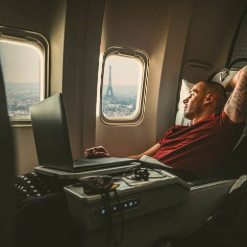 Casually dressed middle aged man working on laptop in aircraft cabin during his business travel, while looking through window at  landmark Eiffel tower.